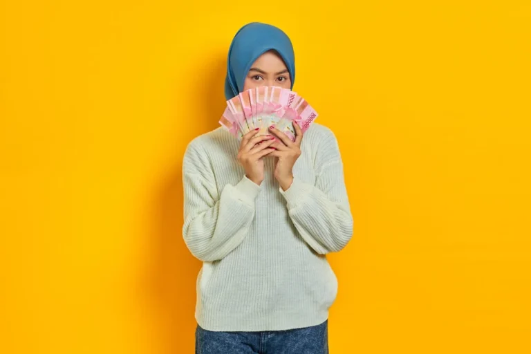 how to withdraw money from binance - Beautiful Asian Muslim woman in white sweater covering face with cash money in Indonesian rupiah banknotes isolated over yellow background. ​People religious lifestyle concept